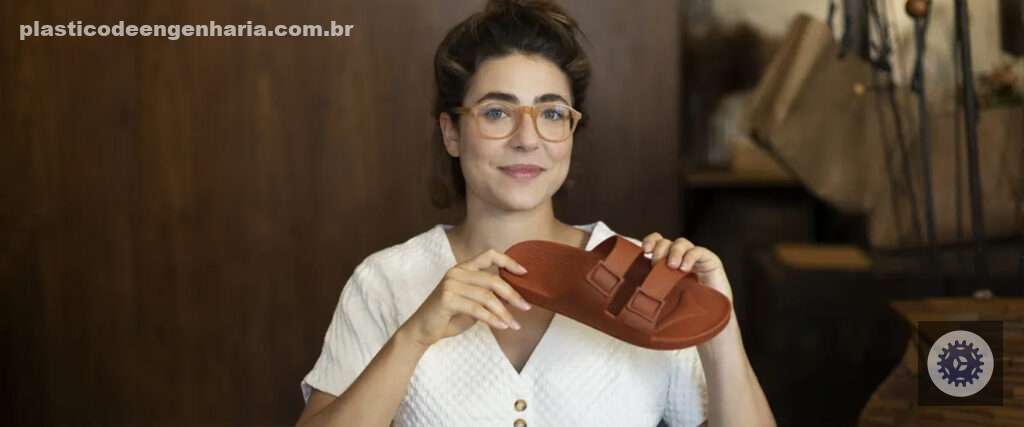 Isabela Chusid premiada por lançar a primeira sandália de plástico vegana. Ela é branca, usa óculos marrom claro, veste uma blusa branca. A sandália em sua mão é da cor marrom.