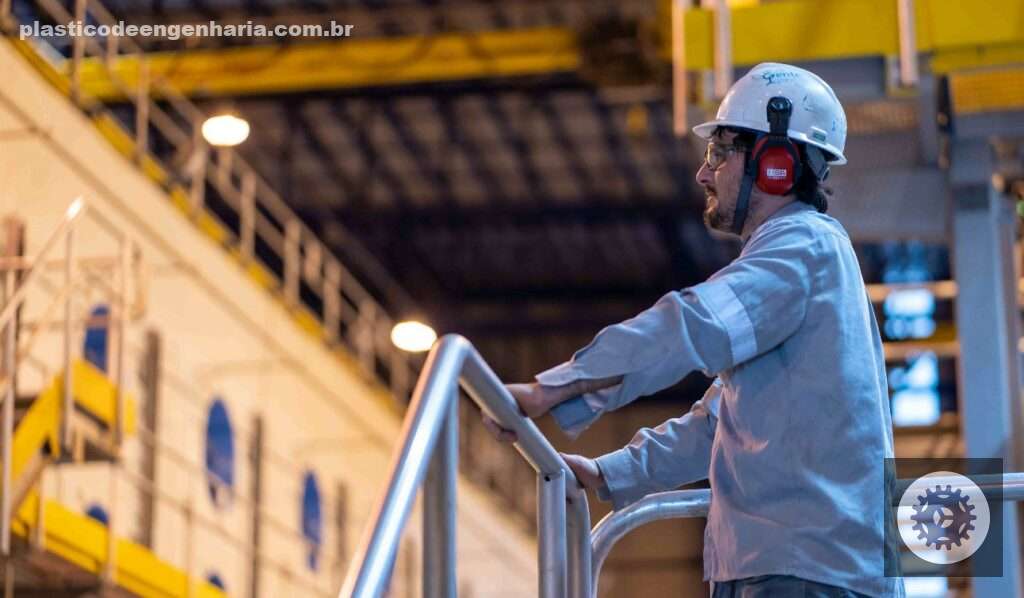 Na imagem aparece um homem com camisa social, ele tem pele branca e barba preta, também usa um capacete e um fone de proteção. ele está em uma indústria. A imagem representa o aumento dos custos industriais
