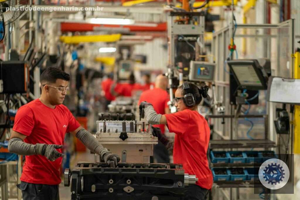 Na imagem aparecem duas pessoas, um homem e uma mulher de pele parda, trabalhando em uma indústria. Eles vestem camisa vermelha, usam óculos de proteção, e luvas cinzas. A imagem representa a confiança industrial de março.